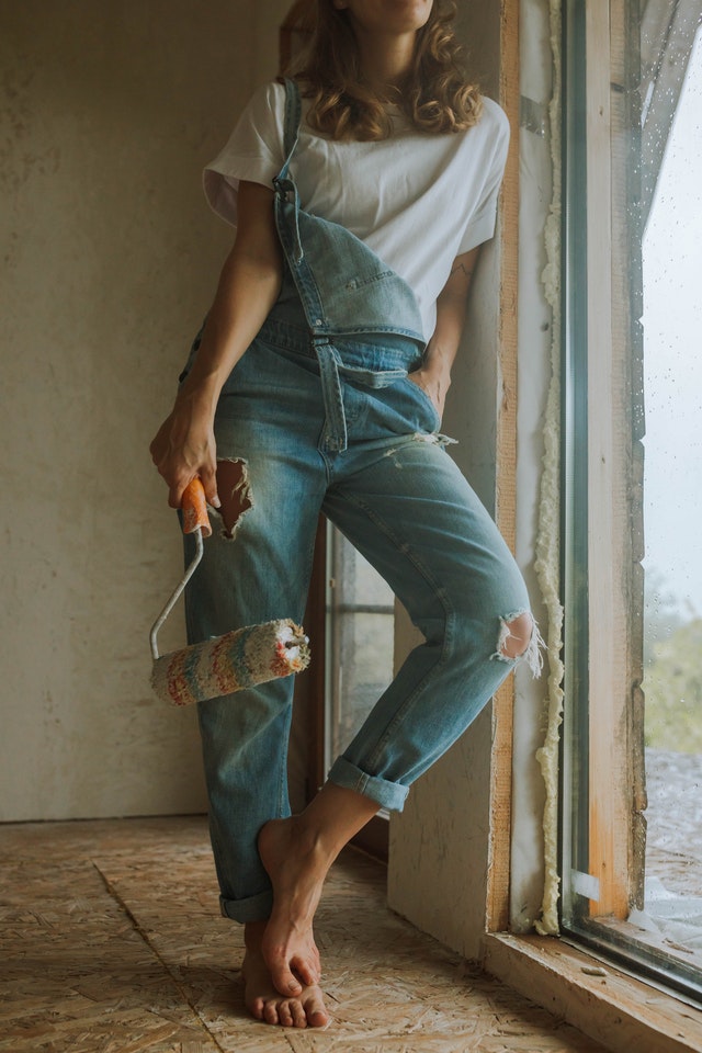 A woman leaning against a window with a paintbrush on a bare floor wearing overalls holding a paintbrush